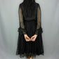 A woman viewed from behind wearing a black midi dress with sheer long sleeves, a ruffled waist, and a black polka-dot tulle skirt, standing against a light gray studio backdrop.