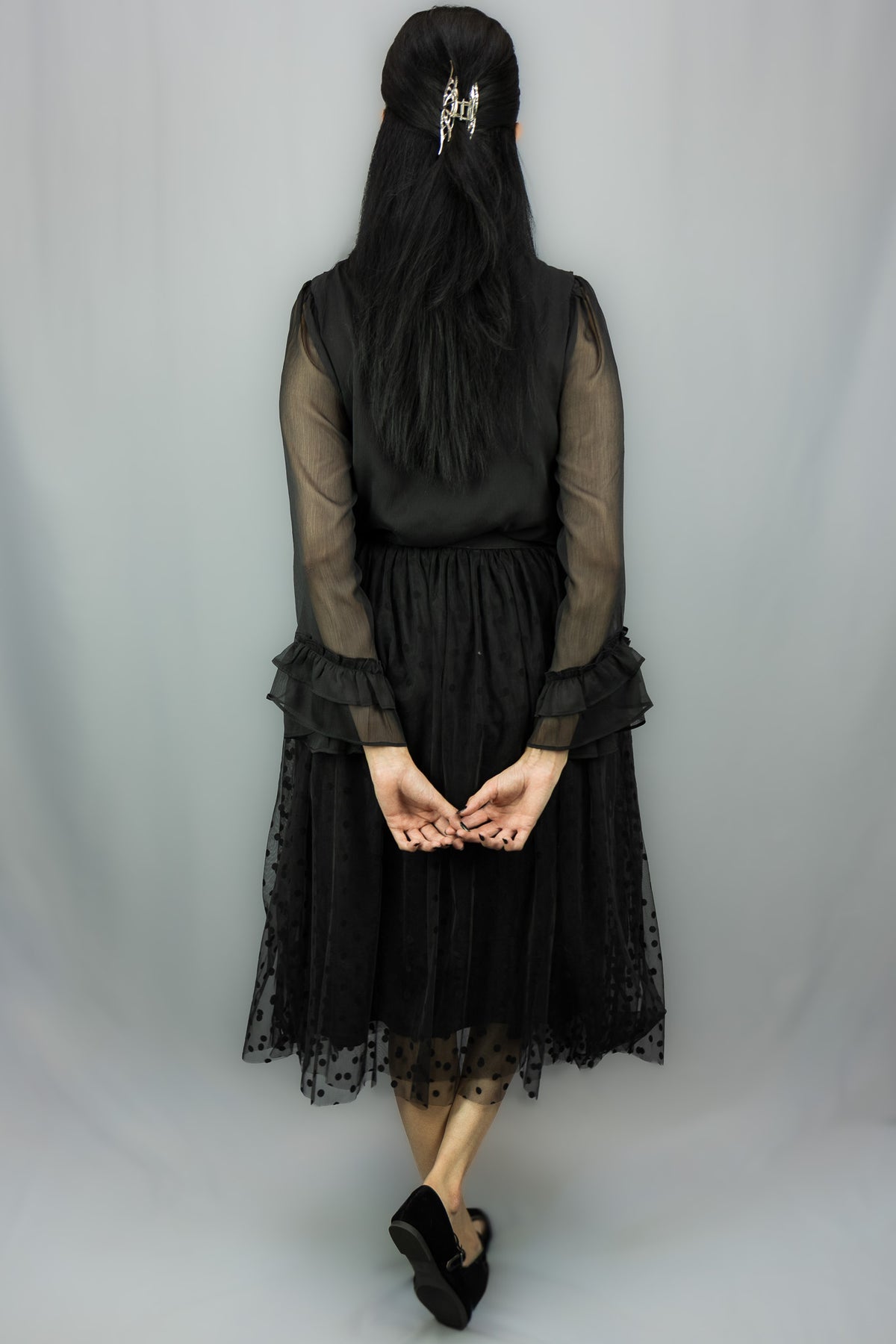 A woman viewed from behind wearing a black midi dress with sheer long sleeves, a ruffled waist, and a black polka-dot tulle skirt, standing against a light gray studio backdrop.