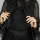 A person wearing a black sheer long-sleeve blouse with a large bow at the neck and a black gathered skirt, holding the sides of the blouse open against a plain light gray background.
