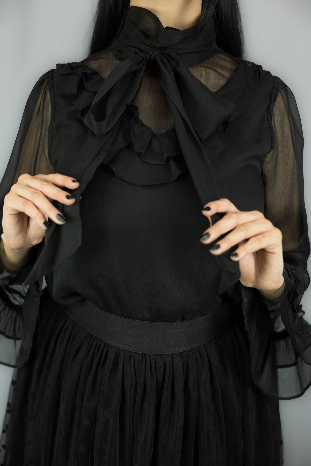 A person wearing a black sheer long-sleeve blouse with a large bow at the neck and a black gathered skirt, holding the sides of the blouse open against a plain light gray background.