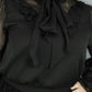 A close-up of a person wearing a black long-sleeve blouse with a sheer ruffled collar, a large front bow, and a black skirt, against a plain gray background.