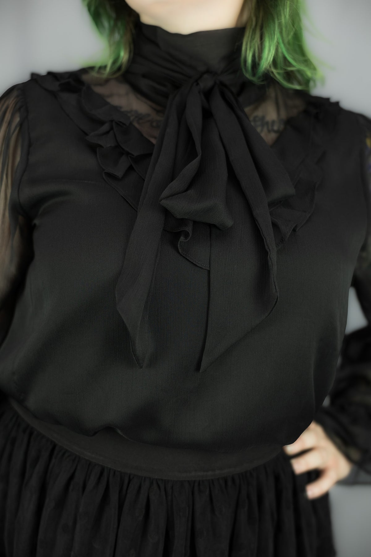 A close-up of a person wearing a black long-sleeve blouse with a sheer ruffled collar, a large front bow, and a black skirt, against a plain gray background.
