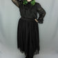 A person modeling a black sheer long-sleeve blouse with a large bow at the neck and a matching black gathered midi skirt with lace trim, paired with black lace-up boots, against a light gray studio backdrop.