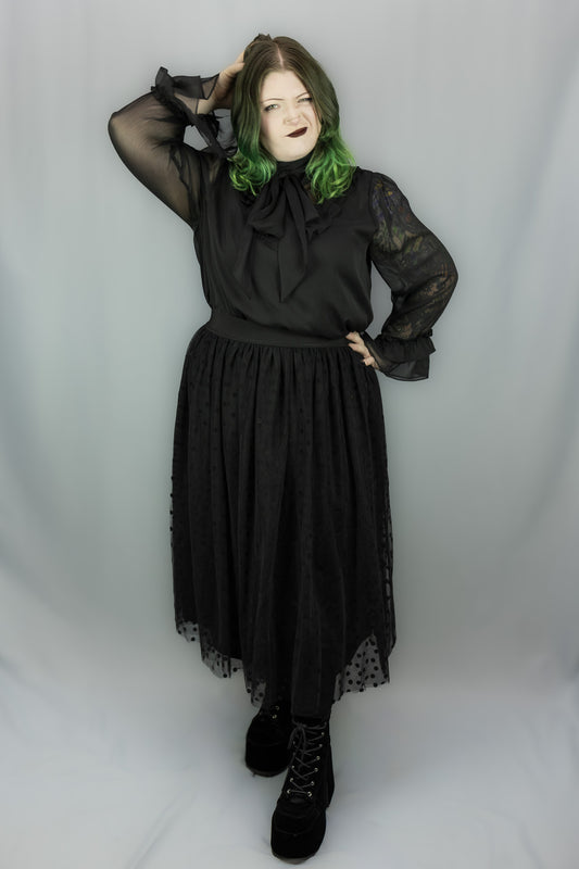 A person modeling a black sheer long-sleeve blouse with a large bow at the neck and a matching black gathered midi skirt with lace trim, paired with black lace-up boots, against a light gray studio backdrop.
