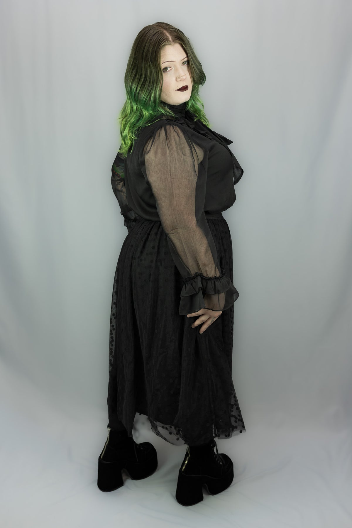 A model wearing a black sheer long-sleeve midi dress with a ruffled collar, paired with black platform boots, standing against a light gray fabric backdrop.