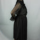 A woman modeling a black sheer long-sleeve blouse with a large bow at the neckline, paired with a black midi skirt featuring a subtle polka-dot pattern and a sheer tulle hem, against a light gray studio background.