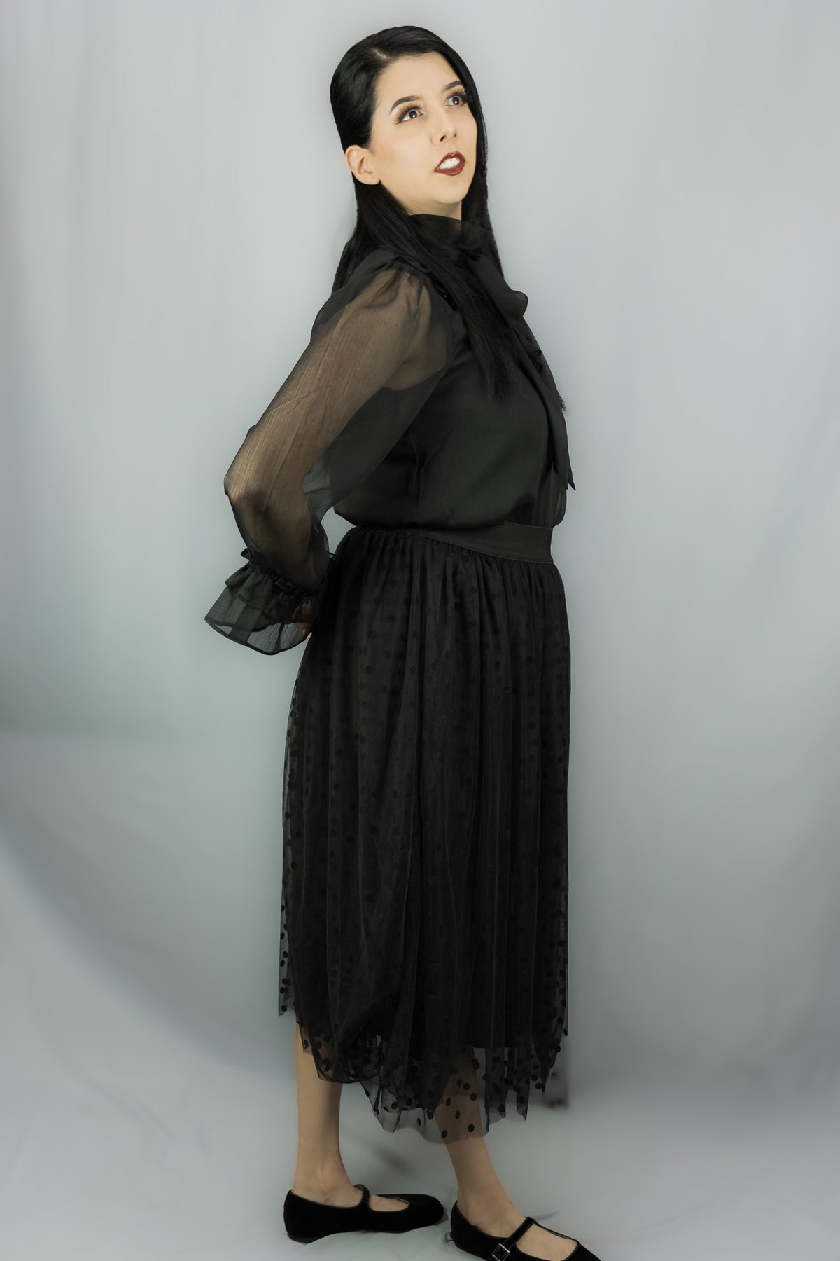 A woman modeling a black sheer long-sleeve blouse with a large bow at the neckline, paired with a black midi skirt featuring a subtle polka-dot pattern and a sheer tulle hem, against a light gray studio background.