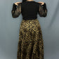 A woman shown from the back wearing a black long-sleeve top with sheer mesh sleeves and a high-waisted leopard-print maxi skirt, standing against a gray studio backdrop.