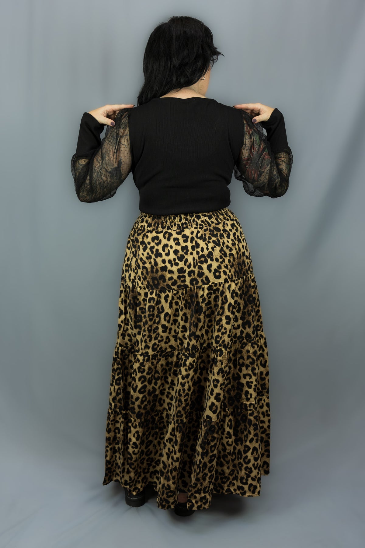 A woman shown from the back wearing a black long-sleeve top with sheer mesh sleeves and a high-waisted leopard-print maxi skirt, standing against a gray studio backdrop.