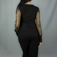 A woman viewed from the back wearing a black long-sleeve top with sheer polka dot mesh sleeves and black wide-leg pants, standing against a light gray studio backdrop.