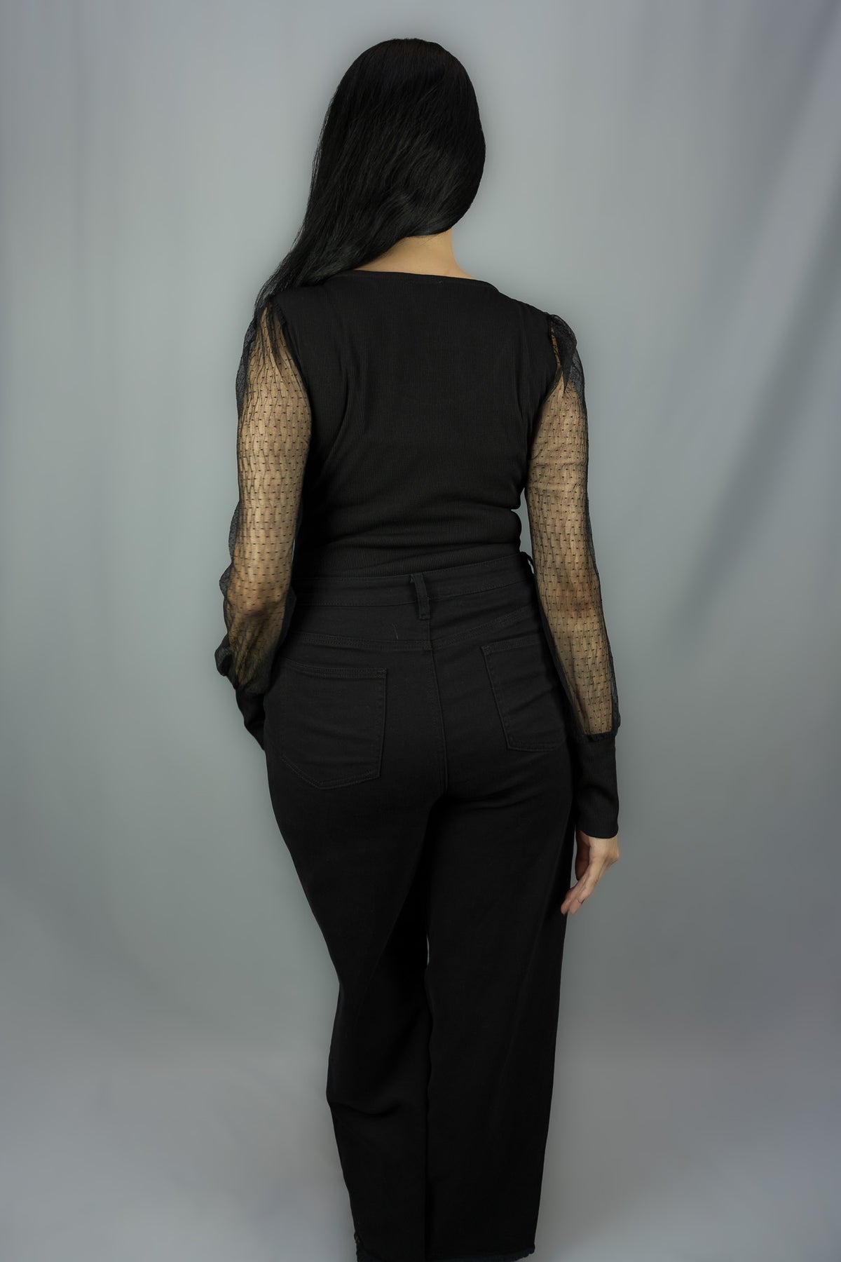 A woman viewed from the back wearing a black long-sleeve top with sheer polka dot mesh sleeves and black wide-leg pants, standing against a light gray studio backdrop.