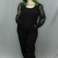 A person with green-tinted hair stands against a light gray studio backdrop wearing a black outfit with sheer lace long sleeves, a black top, black pants, and black shoes.