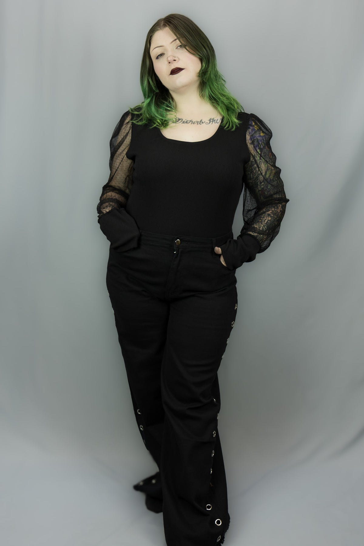 A person with green-tinted hair stands against a light gray studio backdrop wearing a black outfit with sheer lace long sleeves, a black top, black pants, and black shoes.