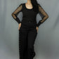 A woman modeling a black outfit consisting of a black long-sleeve top with sheer mesh sleeves and black wide-leg pants with silver eyelet lace-up details down the sides, standing against a gray studio backdrop.
