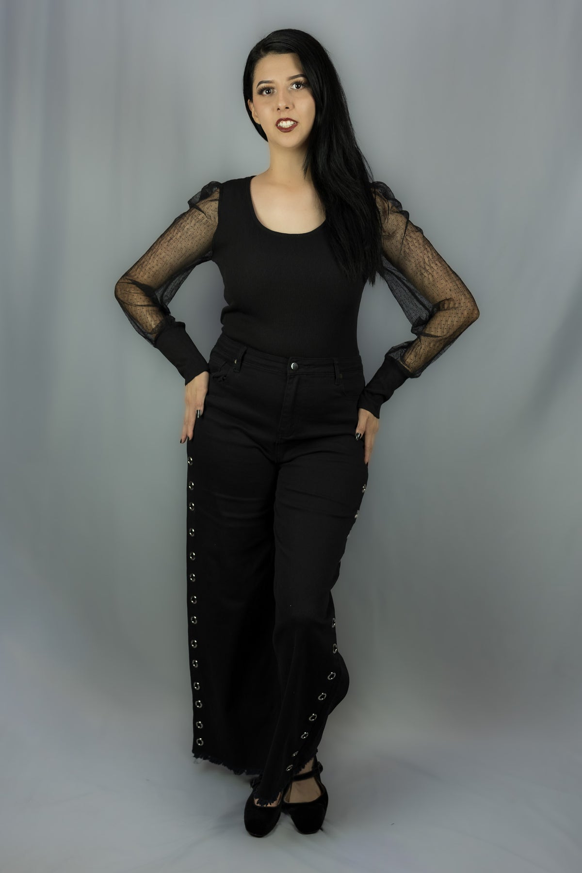 A woman modeling a black outfit consisting of a black long-sleeve top with sheer mesh sleeves and black wide-leg pants with silver eyelet lace-up details down the sides, standing against a gray studio backdrop.