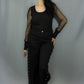 A woman modeling a black gothic-style outfit consisting of a black long-sleeve top with sheer mesh sleeves, black pants with lace-up side details, and black flats, set against a plain gray studio backdrop.