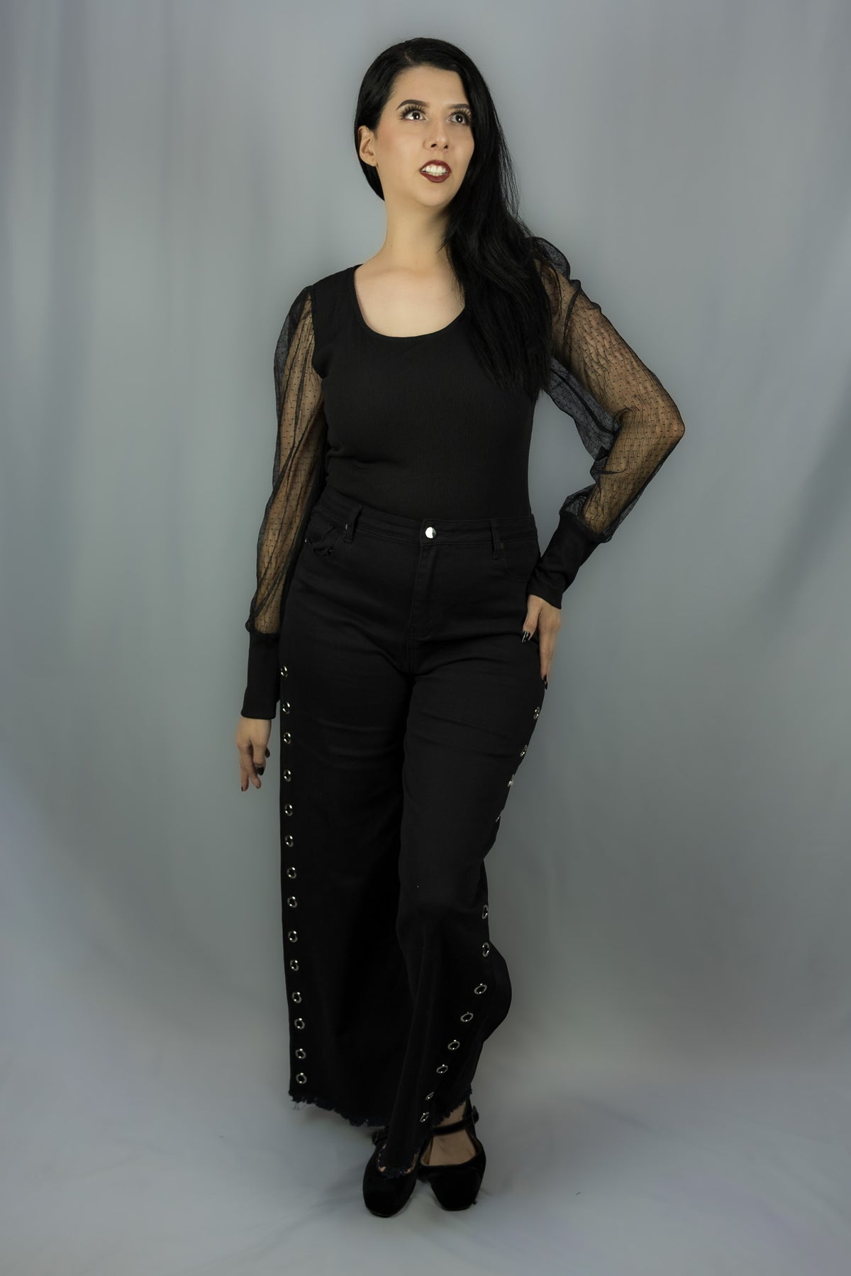 A woman modeling a black gothic-style outfit consisting of a black long-sleeve top with sheer mesh sleeves, black pants with lace-up side details, and black flats, set against a plain gray studio backdrop.