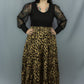 A woman modeling a black long-sleeve top with sheer patterned sleeves and a high-waisted leopard-print maxi skirt, standing against a gray studio backdrop.