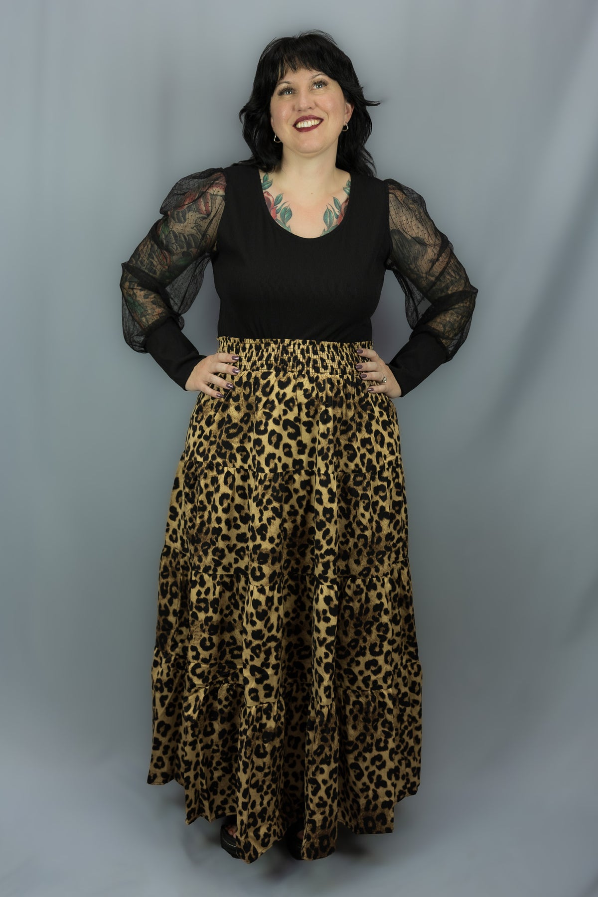 A woman modeling a black long-sleeve top with sheer patterned sleeves and a high-waisted leopard-print maxi skirt, standing against a gray studio backdrop.