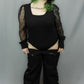A person modeling a black bodysuit with sheer long sleeves and black wide-leg pants against a plain gray studio backdrop.