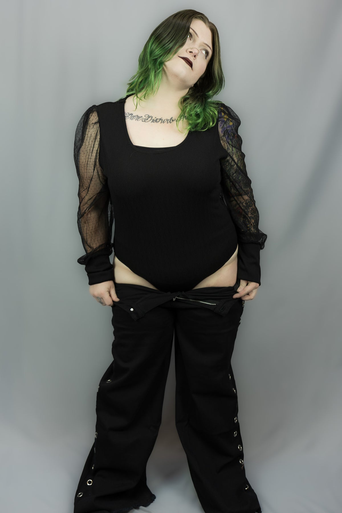 A person modeling a black bodysuit with sheer long sleeves and black wide-leg pants against a plain gray studio backdrop.