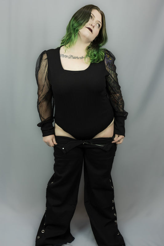 A person modeling a black bodysuit with sheer long sleeves and black wide-leg pants against a plain gray studio backdrop.