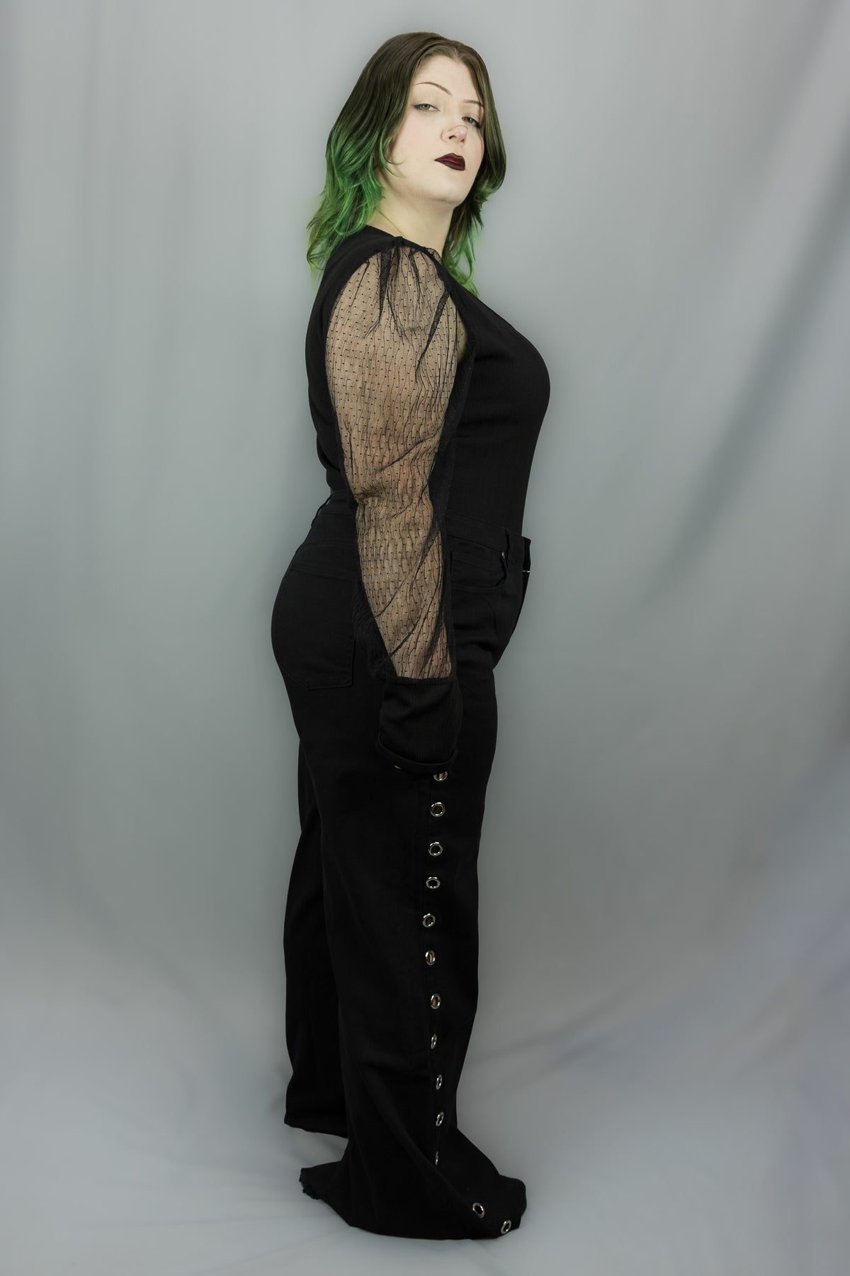 A model wearing a black gothic-style jumpsuit with sheer fishnet mesh sleeves standing against a light gray studio backdrop.
