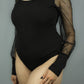 A black long-sleeve bodysuit with sheer mesh sleeves, shown on a model against a plain gray background.