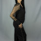 A woman modeling a black outfit with sheer long sleeves and side grommet details, standing against a light gray studio backdrop.