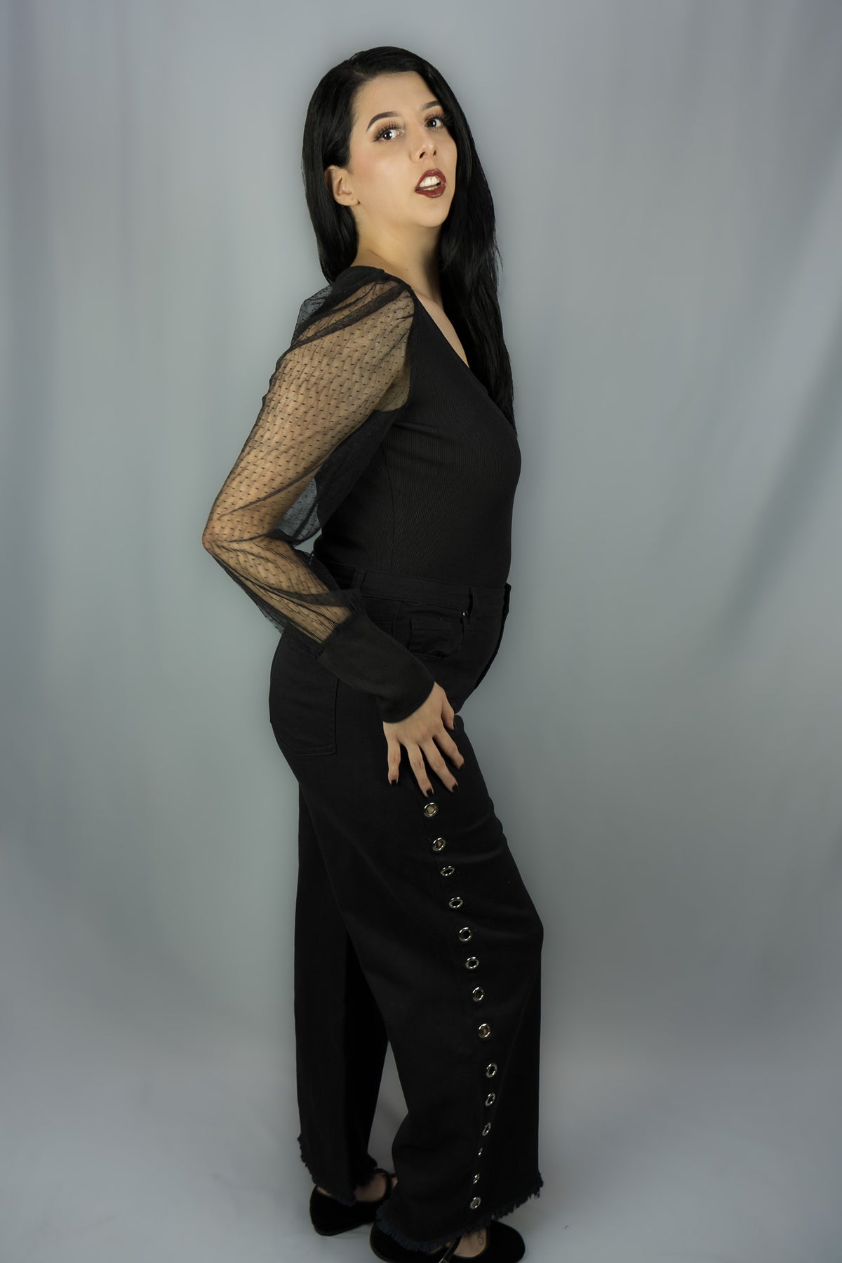 A woman modeling a black outfit with sheer long sleeves and side grommet details, standing against a light gray studio backdrop.