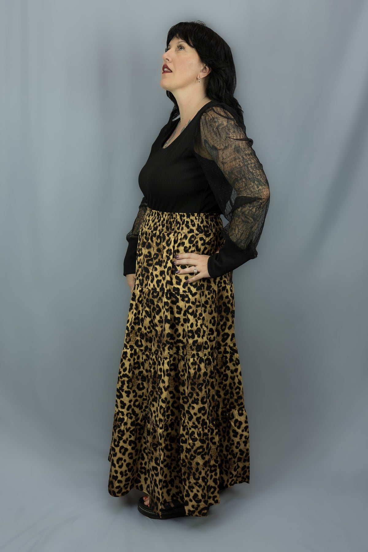 A woman modeling a leopard-print maxi skirt paired with a black long-sleeve top, standing against a gray studio backdrop.
