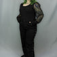 A model with green-tinted hair wearing a black long-sleeve top with sheer polka dot patterned sleeves and black wide-leg pants with grommet details, standing against a gray studio backdrop.