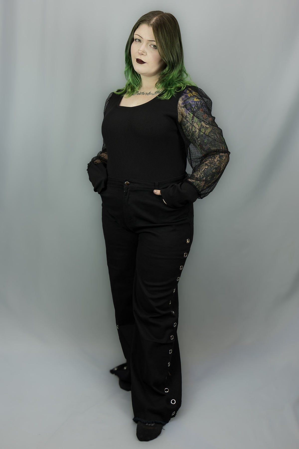 A model with green-tinted hair wearing a black long-sleeve top with sheer polka dot patterned sleeves and black wide-leg pants with grommet details, standing against a gray studio backdrop.