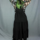 A model viewed from behind wearing a black tiered midi dress over a white and black plaid long-sleeve shirt, with bright green hair, standing against a gray fabric backdrop.