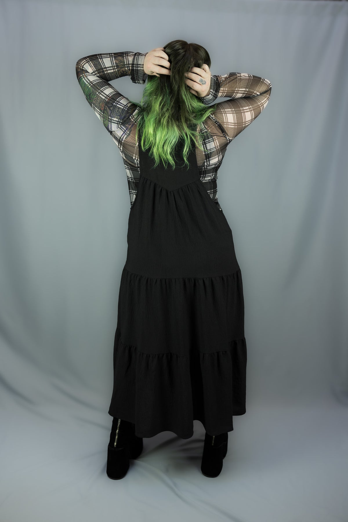 A model viewed from behind wearing a black tiered midi dress over a white and black plaid long-sleeve shirt, with bright green hair, standing against a gray fabric backdrop.
