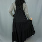 A woman shown from behind wearing a black tiered jumper dress over a black and white striped long-sleeve shirt, standing against a light gray fabric backdrop.
