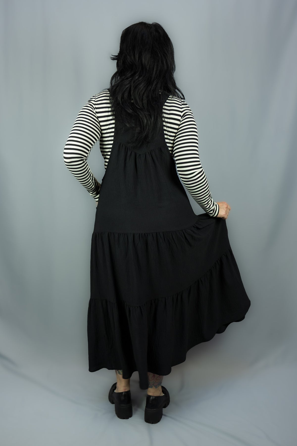 A woman shown from behind wearing a black tiered jumper dress over a black and white striped long-sleeve shirt, standing against a light gray fabric backdrop.