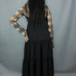 A model shown from the back wearing a black tiered midi dress over a black and white plaid long-sleeve shirt, standing against a gray studio backdrop.