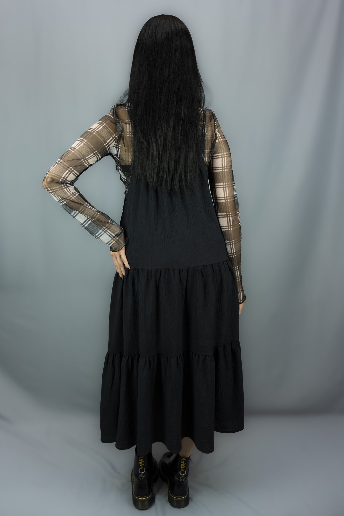 A model shown from the back wearing a black tiered midi dress over a black and white plaid long-sleeve shirt, standing against a gray studio backdrop.