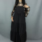 A model with long black hair stands against a gray studio backdrop wearing a black tiered jumper dress over a sheer black and white plaid long-sleeve top and black shoes.