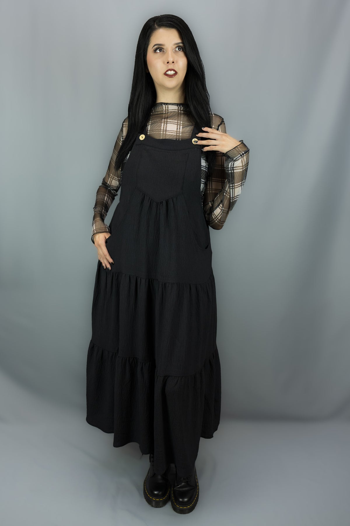 A model with long black hair stands against a gray studio backdrop wearing a black tiered jumper dress over a sheer black and white plaid long-sleeve top and black shoes.