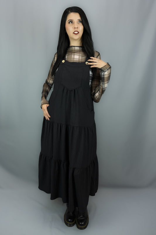 A model with long black hair stands against a gray studio backdrop wearing a black tiered jumper dress over a sheer black and white plaid long-sleeve top and black shoes.