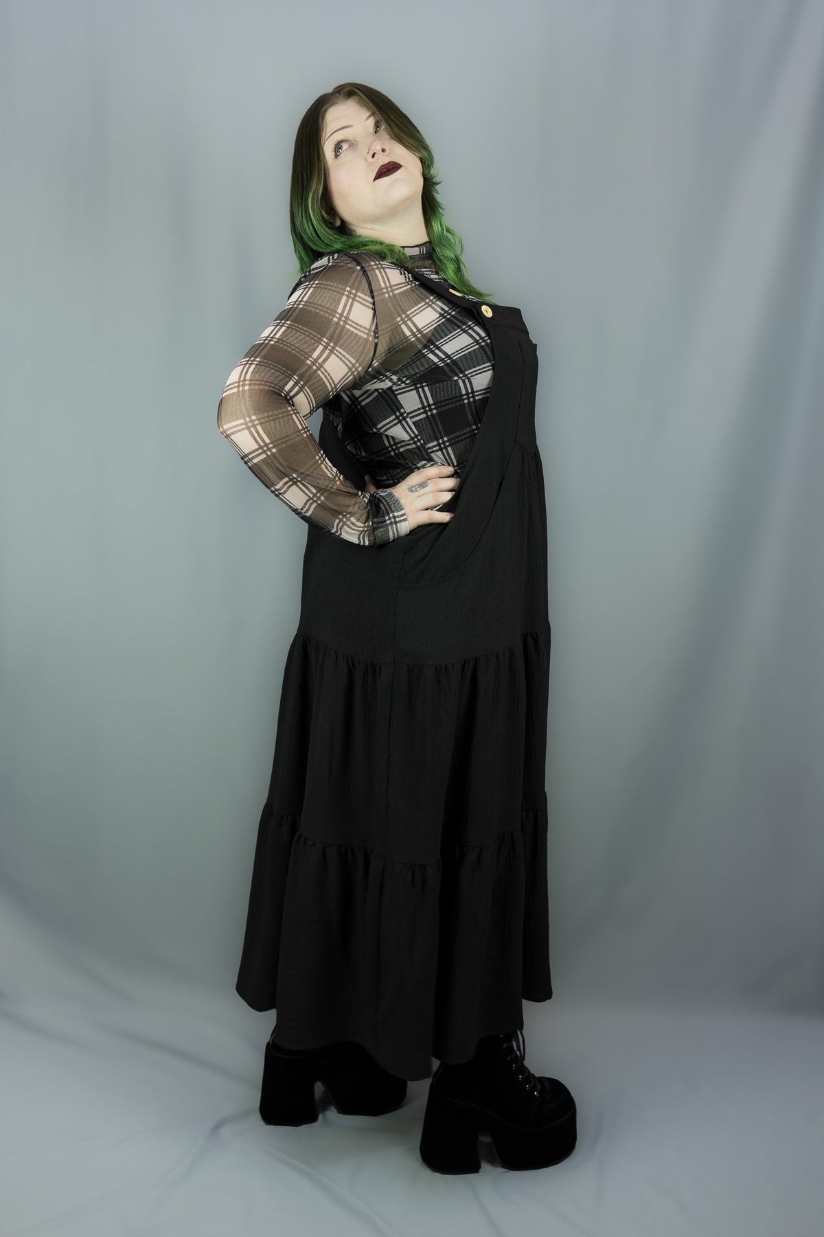 A model wearing a black tiered jumper dress over a sheer black-and-white plaid long-sleeve shirt, styled with black platform boots, against a gray studio backdrop.