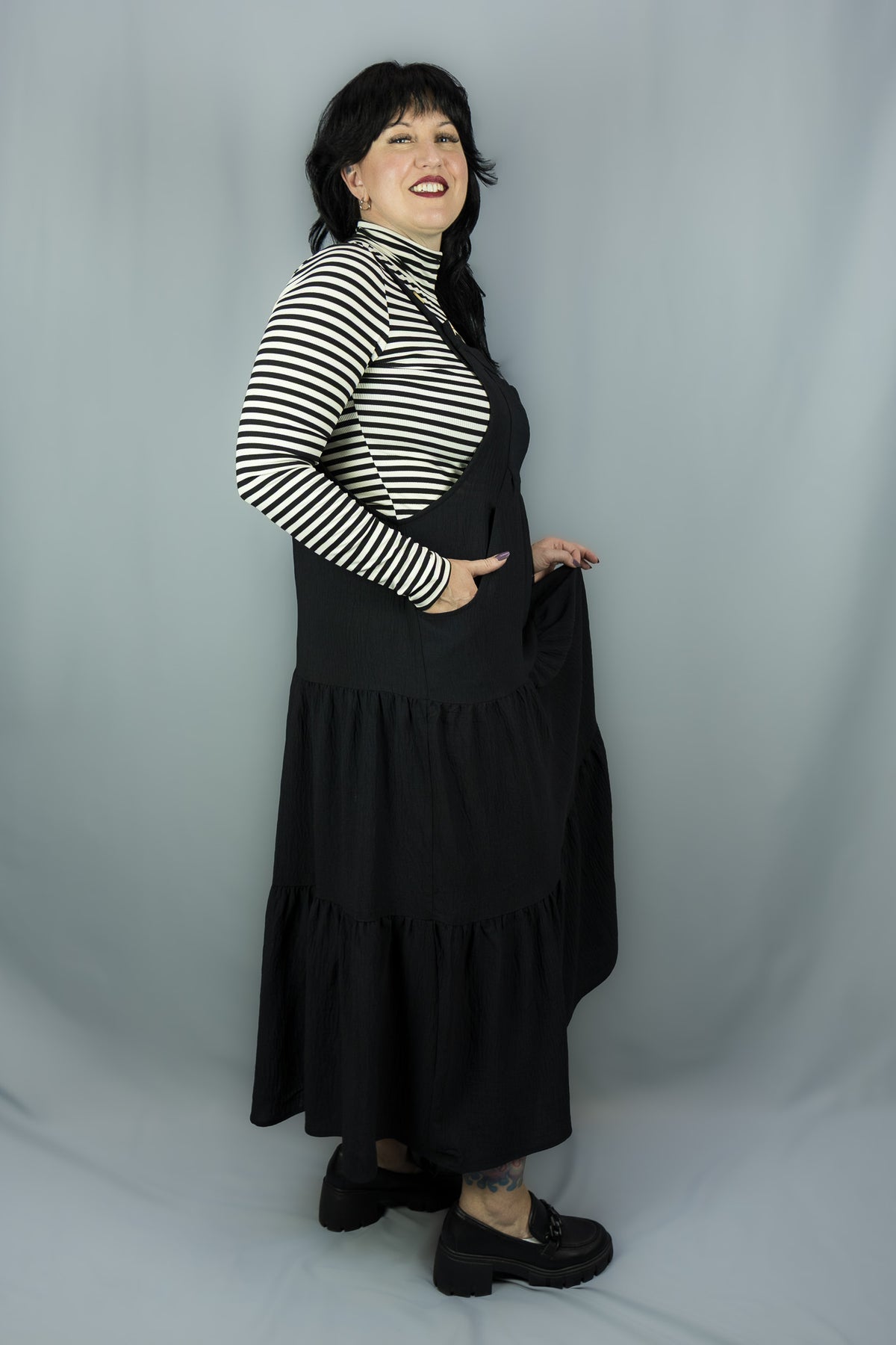 A woman modeling a black tiered jumper dress over a black and white striped long-sleeve turtleneck, standing against a light gray studio backdrop.