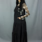 A woman modeling a black tiered midi dress with a sheer black and white plaid long-sleeve shirt underneath, paired with black combat boots, against a gray studio backdrop.