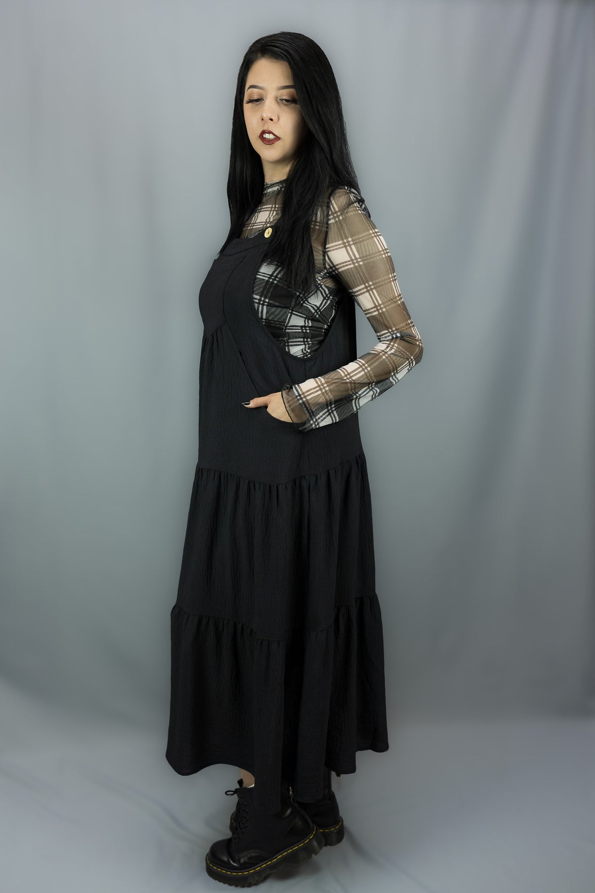 A woman modeling a black tiered midi dress with a sheer black and white plaid long-sleeve shirt underneath, paired with black combat boots, against a gray studio backdrop.