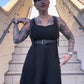 A person wearing a black dress and showcasing a leather fashion harness with adjustable straps and a metal buckle. The harness is designed to be worn around the waist.
