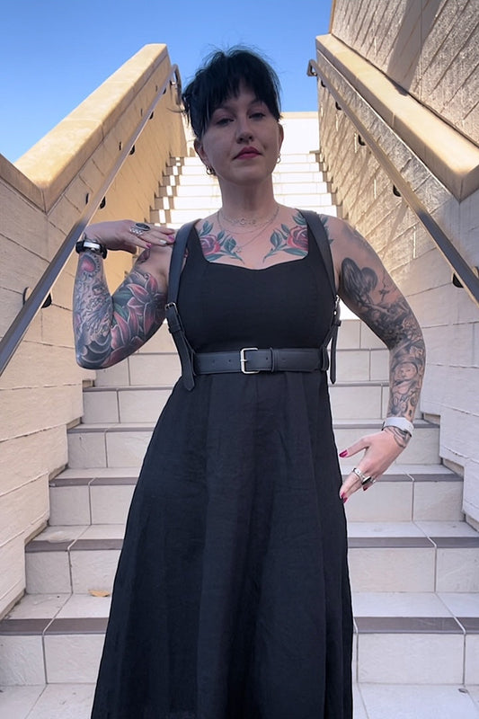 A person wearing a black dress and showcasing a leather fashion harness with adjustable straps and a metal buckle. The harness is designed to be worn around the waist.