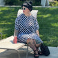 Woman sitting on a chair outdoors, wearing a checkered dress and sunglasses.