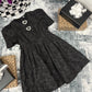 Black dress with decorative buttons on a textured surface with gift boxes and a plant.